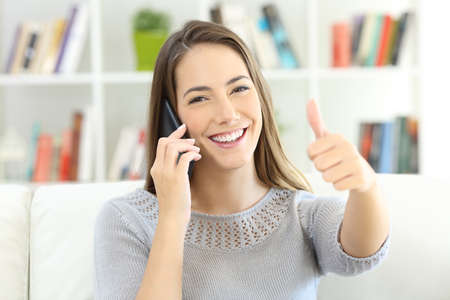 Satisfied customer talking on phone and looking at you sitting on a sofa at homeの写真素材