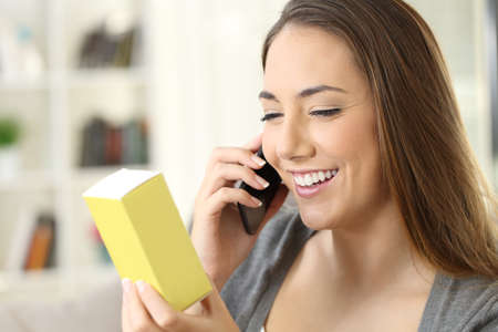 Happy woman asking information on phone about a product sitting on a sofa in the living room in a house interiorの写真素材
