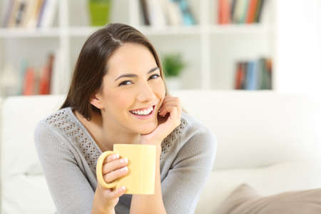 Happy woman posing looking art camera holding a coffee cup sitting on a couch at homeの写真素材
