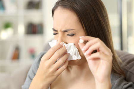 Woman coughing covering mouth with a tissue and holding a pill sitting on a sofa in a house interior