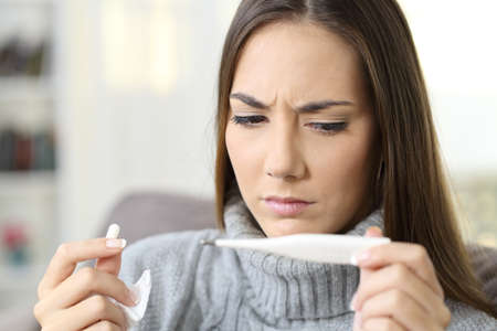 Girl holding a pill and checking thermometer temperature sitting on a sofa in the living room in a house interiorの写真素材