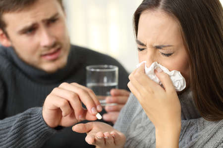 Husband helping his ill wife giving medicines sitting on a couch in a house interiorの写真素材
