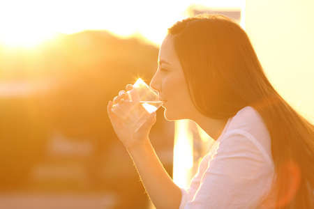 Side view backlight portrait of a woman drinking water from a glass in a house balcony at sunsetの写真素材