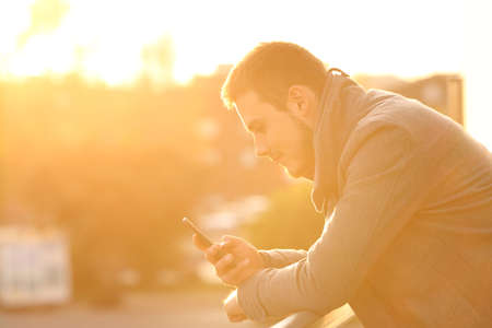 Side view portrait of a man using a smart phone in winter in a house balcony at sunset