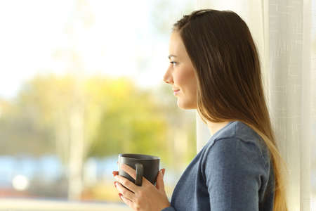 Side view portrait of a serious pensive woman looking through a window at homeの写真素材