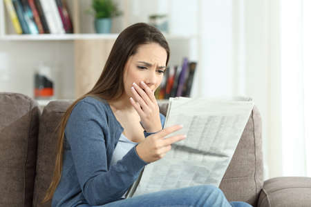 Worried woman reading bad news in a newspaper sitting on a couch in the living room at homeの写真素材
