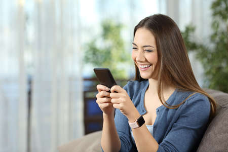 Happy woman wearing a smartwatch using a mobile phone sitting on a couch in the living room at homeの写真素材