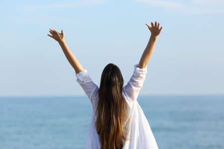 Back view portrait of a happy woman raising arms towards the sea on the beach