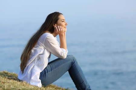 Side view portrait of a happy woman relaxing sitting on the grass watching the sea in the background