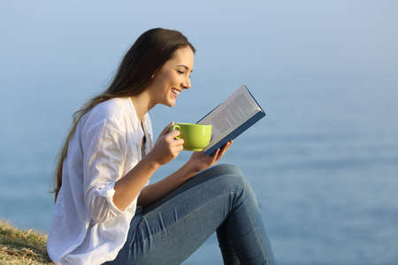 Side view portrait of a happy girl relaxing reading a book and drinking coffee sitting on the grass on the beachの写真素材