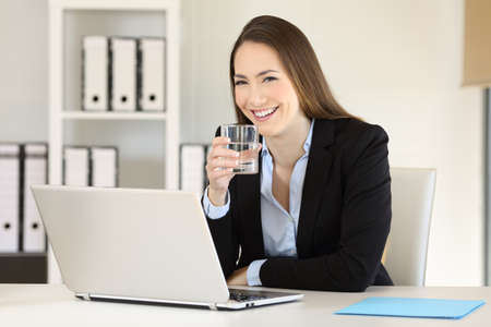 Happy executive holding a glass of water and looking at camera at officeの写真素材