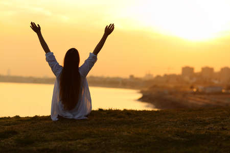 Back view backlight portrait of a happy single woman seeing the city at sunset and raising arms with a warm light in the backgroundの写真素材