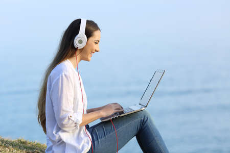 Side view portrait of a happy woman watching and listening online content in a laptop outdoors on the beachの写真素材