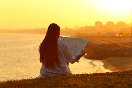 Back view backlight portrait of a tourist reading a map at sunset with a beautiful landscape in the backgroundの写真素材