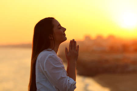 Side view backlight portrait of a woman praying and looking above at sunsetの写真素材