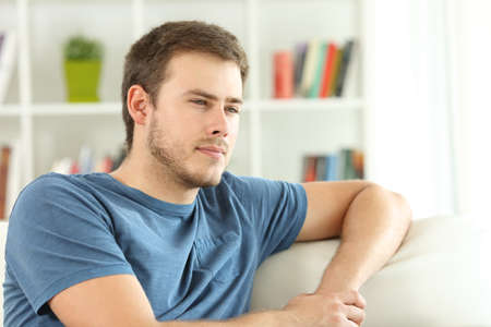 Serious man thinking looking away sitting on a couch in the living room at homeの写真素材