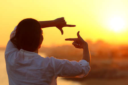 Back view backlight portrait of a woman framing with fingers at sunsetの写真素材