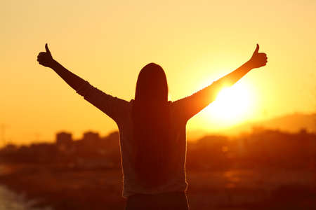 Back view backlight silhouette of a woman raising arms with thumbs up to the sun at sunset