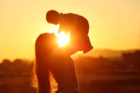 Backlight silhouette of a mother raising her kid son at sunset with the sun in the middleの写真素材