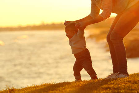 Backlight silhouette of a confident baby learning to walk and his mom helping him at sunsetの写真素材