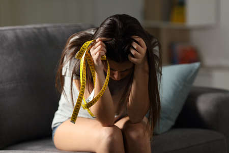 Sad teen worried about appearance holding a measure tape sitting on a couch in the living room at homeの写真素材