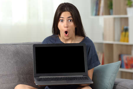 Amazed teen showing you a blank laptop screen sitting on a couch in the living room at homeの写真素材