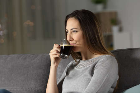 Relaxed woman drinking decaffeinated coffee in the night sitting on a couch in the living room at...