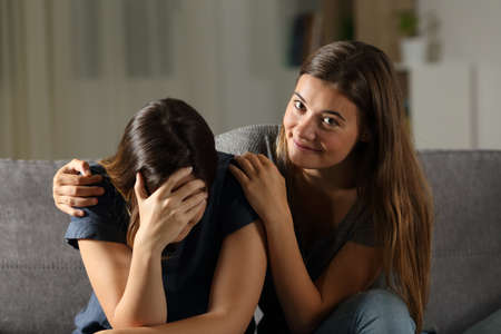 Bad teen is happy with her sad friend crying sitting on a couch in the living room at home