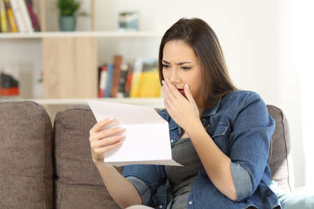 Sad woman reading bad news in a paper letter sitting on a couch in the living room at homeの写真素材