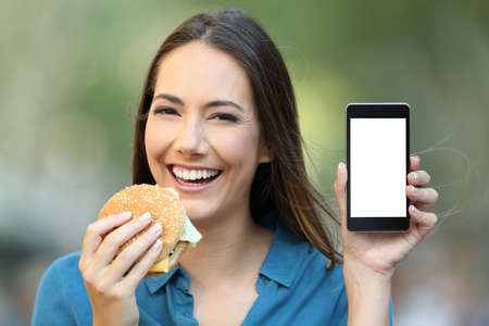 Happy woman holding a hamburger showing a phone screen in the street