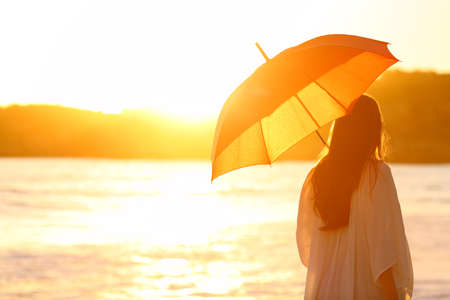 Back view portrait of a woman walking with an umbrella at sunset on the beachの写真素材