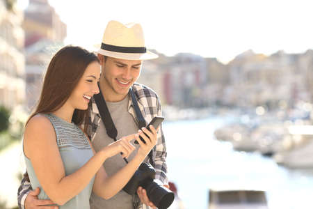 Couple of happy tourists booking hotel online with a smart phone on vacation in a coast town