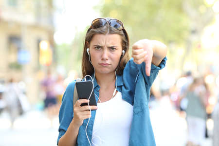 Front view portrait of an annoyed girl listening to music with thumbs down in the streetの写真素材