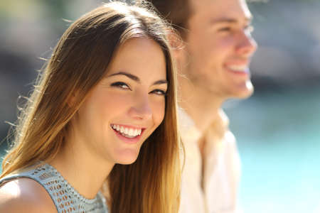 Happy woman smiling with perfect teeth looking at you beside her partner on the beachの写真素材