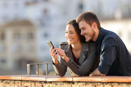 Happy couple using smart phone in a balcony at sunset with a town in the background