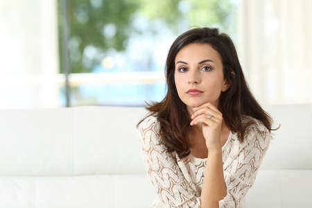 Front view of a confident serious woman looking at camera sitting on a couch in the living room at...
