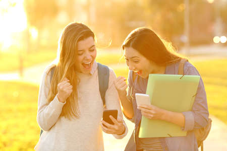 Two excited students finding good news online in a smar phone in a park at sunsetの写真素材