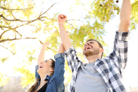 Excited couple celebrating success raising arms in a park with trees in the background