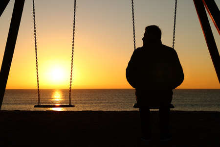 Back view backlighting silhouette of a man alone on a swing looking at empty seat at sunset on the beach in winterの写真素材
