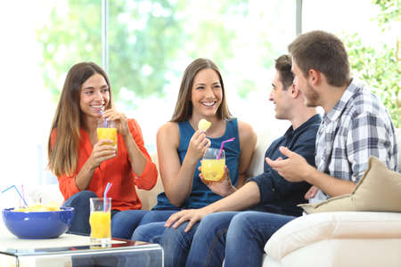 Group of happy friends talking eating chips and drinking refreshments sitting on a couch in the living room at homeの写真素材