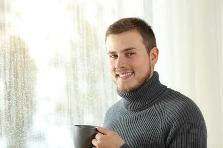 Happy tenant looking at camera beside a window at home a rainy day