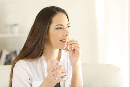 Happy woman taking vitamin pill holding glass sitting on a couch at homeの写真素材