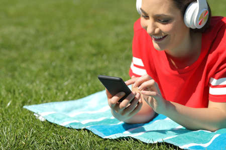 Happy girl in red listens to music browsing smart phone content on the grass