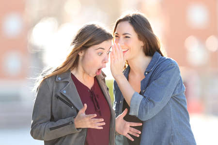 Two gossip girls whispering on ear a secret standing in the street