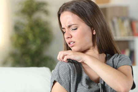 Woman suffering itching scratching shoulder sitting on a couch at home