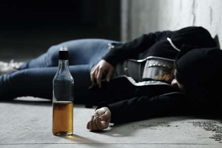 Drunk girl sleeping on the floor in a garage with a bottle of alcohol drink in foreground
