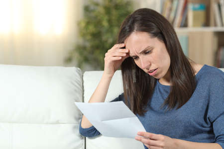 Worried woman reading a letter sitting on a couch in the living room at homeの写真素材