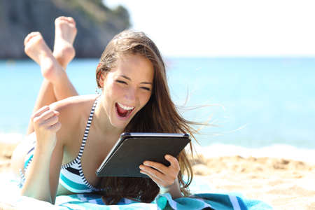 Excited girl in bikini checking tablet content lying on the sand on the beach on summer vacationの写真素材