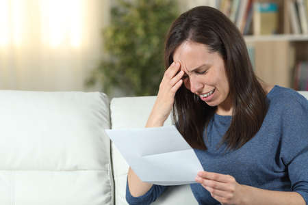 Sad woman reading a letter sitting on a couch in the living room at homeの写真素材