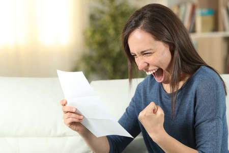 Excited woman reading a letter sitting on a couch in the living room at homeの写真素材